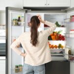 Young woman near open refrigerator in kitchen, back view